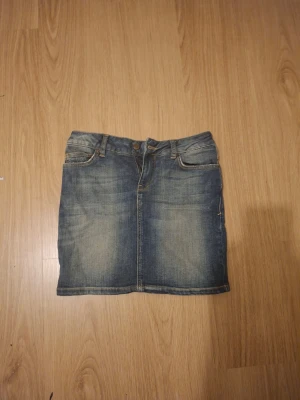 Kort jeanskjol i blå denim - Snygg kort jeanskjol i klassisk blå denim med femficksmodell och knappgylf. Kjolen har en rak passform och slitna detaljer framtill för en avslappnad look. Perfekt att matcha med sneakers eller boots.