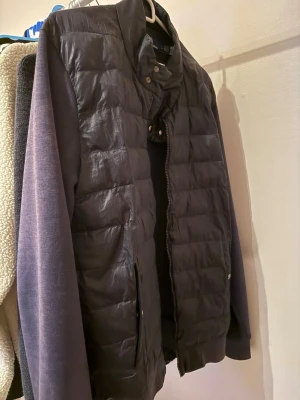 Svart dunväst från Polo Ralph Lauren - Snygg svart dunväst från Polo Ralph Lauren med quiltad design och hög krage. Västen har dragkedja framtill, tryckknappar vid kragen och två fickor med knappstängning. Perfekt att slänga över en hoodie för en chill look. Passformen är L/XL, samt den är solblekt 