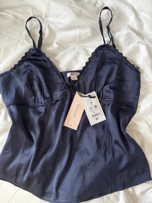 Mörkblått satinset från Gina Tricot - Säljer ett mörkblått satinset från Gina Tricot med ett linne och matchande shorts. Linnet har tunna axelband och spetsdetaljer vid ringningen. Shortsen har elastisk midja och volangkant nedtill.🩷aldrig använda med prislapp kvar, storleken är M från Gina Young 