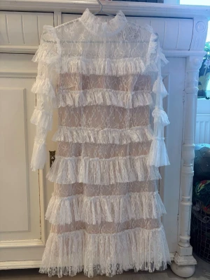 Vit spetsklänning från by malina  - Vit klänning i spets från by malina  flera lager volanger och hög krage. Klänningen är långärmad och har transparent spetsmaterial som ger en elegant look.