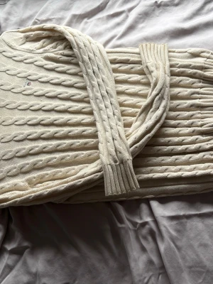 Vit kabelstickad tröja Tommy Hilfiger - Säljer en vit kabelstickad tröja från Tommy Hilfiger med klassiskt flätmönster och diskret logga broderad på bröstet. Tröjan har ribbade muddar och är gjord i mjukt material som känns skönt mot huden. Perfekt för dig som gillar stilrena plagg.