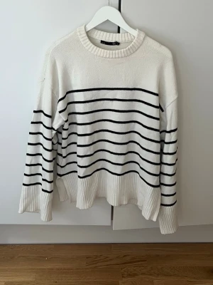 Vit stickad tröja med svarta ränder - Vit stickad tröja med svarta horisontella ränder och rund hals. Tröjan har långa ärmar, ribbstickade muddar och en lös, avslappnad passform. Perfekt för lager-på-lager och enkel att styla till jeans eller kjol. Lätt nopprig.