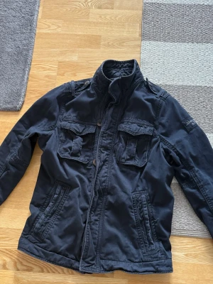 Mörkblå jacka från Abercrombie & Fitch - Snygg mörkblå jacka från Abercrombie & Fitch med hög krage, fyra framfickor och broderad logga på ärmen. Jackan har dragkedja och knappar framtill samt detaljer på axlarna. Perfekt för dig som vill ha en stilren och funktionell jacka. Skriv vid frågor eller funderingar. Är storlek XL men sitter mer som M/L