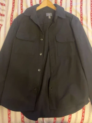 Svart overshirt från H&M - Stilren svart overshirt från H&M med klassisk krage och två stora bröstfickor med lock. Rak passform och knäppning framtill med fyra knappar. Perfekt att bära som lager över t-shirt eller hoodie. Materialet ser ut att vara mjukt och lite tjockare, troligen bomull.