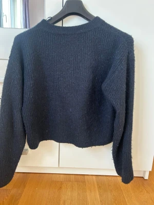 Mörkblå stickad tröja med rosetter - Mysig mörkblå stickad tröja med långa ärmar och rund hals. Baksidan har en unik detalj med tre svarta rosetter som knyts, vilket ger en trendig och personlig touch. Perfekt för dig som gillar stilrena plagg med något extra.