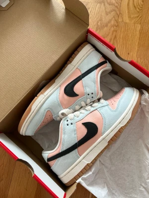 Nike Dunk Low rosa och ljusblå - Nike Dunk Low sneakers i en fräsch färgkombo med ljusrosa och ljusblå mocka, svart swoosh och vit mellansula. Yttersulan är i gummi för extra street-vibe. Klassisk låg siluett och perforerad tåbox för skön ventilation. Perfekta för dig som vill sticka ut med stil.