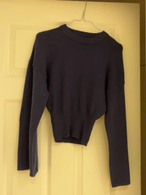 Blå cropped stickad tröja - En blå stickad tröja med cropped passform och rund halsringning. Tröjan har långa ärmar och ribbstickade detaljer vid hals, ärmslut och nederkant. Perfekt för att skapa en avslappnad och trendig look.