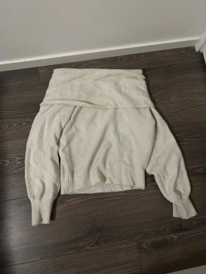 Offshoulder stickad tröja från NewYorker - Krämvit stickad offshoulder-tröja från NewYorker med bred krage och långa ärmar. Tröjan har ribbade muddar vid ärmslut och nederkant, vilket ger en snygg och avslappnad look. Perfekt för dig som gillar trendiga och bekväma plagg. Tröjan är i storlek 38.
