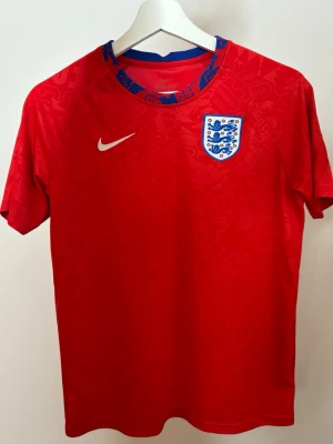England röd matchtröja Nike - Snygg röd England matchtröja från Nike med korta ärmar och rund hals. Tröjan har det klassiska engelska landslagsmärket på bröstet och en vit Nike-logga. Tillverkad i lätt och ventilerande funktionsmaterial, perfekt för fotboll eller träning.