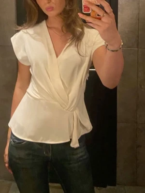 Vit blus med knyt - Stilren vit omlottblus med knyt i sidan och v-ringning. Blusen har korta ärmar och är tillverkad i ett mjukt, lätt material som ger ett snyggt fall. Perfekt att matcha med jeans eller kjol för en trendig look.