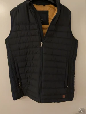 Svart dunväst från Jack & Jones - Stilren svart dunväst från Jack & Jones med gul insida. Västen har dragkedja framtill, två sidofickor med dragkedja och quiltad design. Använd fåtal gånger. Pris går att diskutera 