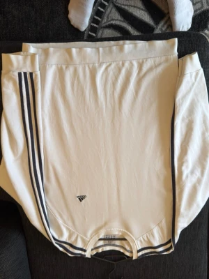 Vit Adidas tröja med svarta ränder - Säljer en vit tröja från Adidas med klassiska svarta ränder längs ärmarna och Adidas-logga framtill. 