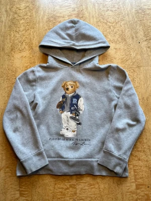 Grå hoodie Polo Bear Ralph Lauren 10-12år - Snygg grå hoodie från Ralph Lauren med den ikoniska Polo Bear-trycket framtill. Mjuk bomullsmix, klassisk huva och ribbade muddar. Perfekt för en avslappnad och trendig look. Trycket visar en nalle i collegejacka och sneakers.