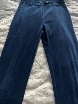 Mörkblå mjukisbyxor i velour - Säljer ett par mörkblå mjukisbyxor i mjuk velour med raka ben och resår i midjan. Byxorna har dragsko med metallöljetter och en avslappnad passform. Perfekta för chill dagar eller när du vill vara extra bekväm.