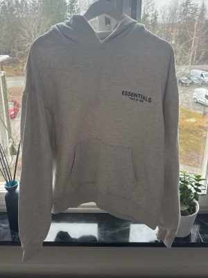 Grå Essentials Fear of God hoodie - Snygg grå hoodie från Essentials Fear of God med svart logga tryckt på bröstet och ryggen. Hoodien har en klassisk känguruficka framtill och en stor huva. Tillverkad i mjukt bomullsmaterial som är perfekt för en avslappnad streetwear-look.