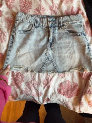 Ljusblå jeanskjol med rå kant - Säljer en ljusblå jeanskjol med rå, fransig kant och slitna detaljer. Kjolen har klassiska fem fickor och snygga bakfickor med broderi. Perfekt för dig som gillar en avslappnad och trendig stil.