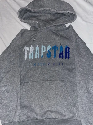 Grå Trapstar hoodie med tryck - Snygg grå hoodie från Trapstar med stor huva och dragsko. Framtill står det 'TRAPSTAR' i olika blå nyanser och 'IT'S A SECRET' spegelvänt under. Klassisk känguruficka och ribbade muddar. Perfekt för en chill och streetwear-inspirerad look. Helt äkta är dom