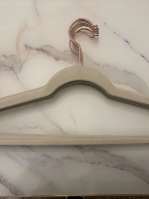 Beige sammetsgalge med roséguldkrok - Snygg beige galge i sammet med krok i roséguld. Perfekt för att hänga upp kläder utan att de glider av. Galgen har en smal och stilren design som sparar plats i garderoben och ger en lyxig känsla.