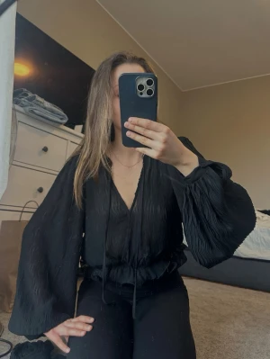 Svart blus med volang  - Snygg svart blus med v-ringning och volangdetalj i midjan. Tyget är tunt och lätt genomskinligt, och den har knappar i ärmarna💕det finns även ett band som man kan sätta runt midjan för en snygg detalj! Helt oanvänd med prislapp kvar🩷