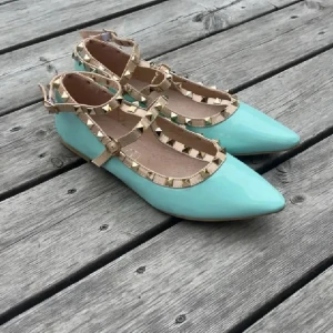 Valentinodupe skor  - Snygga turkosa ballerinaskor med spetsig tå och beigea remmar dekorerade med guldiga nitar. Skorna har en platt sula och stängs med spänne runt vristen. Perfekta för dig som vill sticka ut med en cool detalj och fräsch färg.