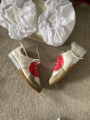 Maison Margiela Japan 2006 Gats sneakers - Den första modellen av Maison Margielas Gats sneakers i vitt läder med beige mockadetaljer och en stor röd cirkel på sidan. Skorna har röda och vita skosnören samt gummisula. Klassisk låg modell med retrovibbar och tydliga kontrastfärger.
