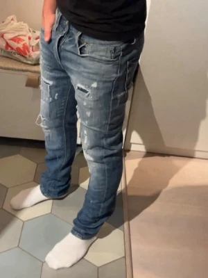 Jack and Jones jeans med slitningar  - Säljer ett par blå jeans med slitningar och hål på benen. Jeansen har en rak passform och klassisk femficksdesign. Materialet är denim och de har en cool, avslappnad look som passar perfekt till en streetwear-stil. Jack and Jones jeans med slitningar || med slitningar || W29 L32 || 