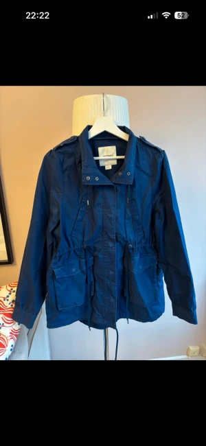 mörkblå knyt jacka  - Blå canvas-jacka från Monki. Går att dra in i midjan beroende på hur insvängd man vill ha den. 100% bomull.  Fint men använt skick. Framför allt visst slitage vid knappar där tyget ljusnat, se bild.