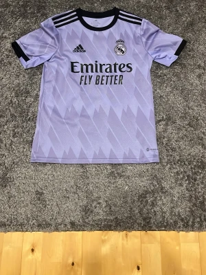 Lila Real Madrid matchtröja Adidas 164 - Snygg lila Real Madrid matchtröja från Adidas med svart krage och svarta detaljer på ärmarna. Tröjan har klubbmärke, Adidas-logga och Emirates Fly Better-tryck på bröstet. Tillverkad i lätt och ventilerande material, perfekt för fotbollsträning.