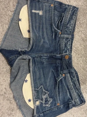 Blå jeansshorts med stjärndetaljer - Säljer ett par blå jeansshorts från HM med råa kanter och slitningar. Shortsen har en cool stjärndetalj på ena benet och innerfoder med blå stjärnor. Klassisk femficksmodell och knappgylf. Perfekta för sommaren! 