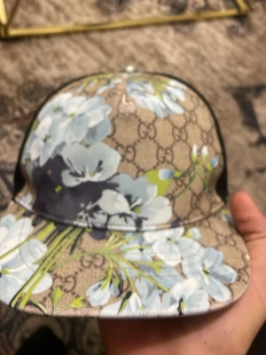  Gucci keps med blommor - Säljer en snygg Gucci keps med klassiskt GG-mönster i beige och brunt, dekorerad med stora blå och vita blommor samt gröna detaljer. Kepsen har svart mesh baktill och justerbar knapp. Perfekt för dig som vill sticka ut med en unik accessoar. Alla skador på kepsen finns i bilderna, kommer inte med lapp. Jag är inte 100% om den är äkta