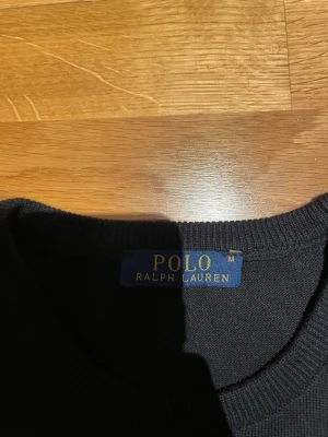 Svart stickad tröja Polo Ralph Lauren - Klassisk svart stickad tröja från Polo Ralph Lauren med korta ärmar och rund hals. Tröjan har den ikoniska röda loggan broderad på bröstet och ribbade muddar vid ärmslut och nederkant. Perfekt för en clean och stilren look.