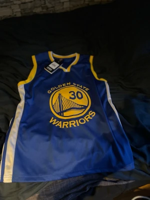 Golden State Warriors Curry tröja - Snygg blå basketlinne med gula och vita detaljer från Golden State Warriors. Framsidan har lagets logga och nummer 30, baksidan har Curry och nummer 30. Perfekt för basketfans! Tillverkad i lätt och ventilerande material.