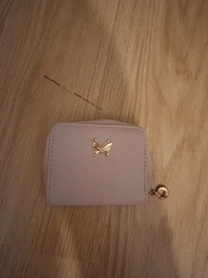 Ljusrosa plånbok med fjäril - Söt ljusrosa plånbok i ett slätt material med guldfärgad fjärilsdetalj på framsidan. Plånboken har dragkedja runtom och en liten guldfärgad ring som detalj. Perfekt storlek för kort och mynt.