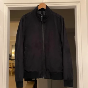 Mörkblå bomberjacka från mango - Stilren mörkblå bomberjacka från MNG med hög krage och dragkedja framtill. Jackan har ribbade muddar vid ärmslut och nederkant samt två sidofickor. Materialet ser ut att vara mjukt och matt, vilket ger en modern look.