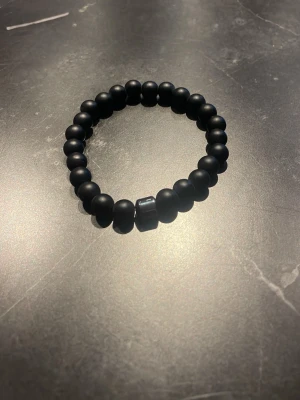 Svart pärlarmband med matt finish - Snyggt svart armband med matta pärlor och en blank, fyrkantig pärla som detalj. Armbandet är elastiskt och passar de flesta handleder. Perfekt accessoar för en stilren och modern look.