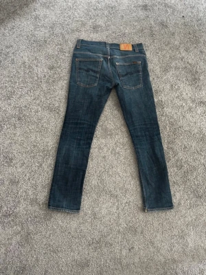 Mörkblå slim jeans från Nudie Jeans - Snygga mörkblå slim fit jeans från Nudie Jeans med klassisk femficksmodell och kontrastsömmar. Jeansen har en skinnpatch med logga bak i midjan och är tillverkade i slitstarkt denimtyg. Perfekta för en avslappnad och trendig look.
