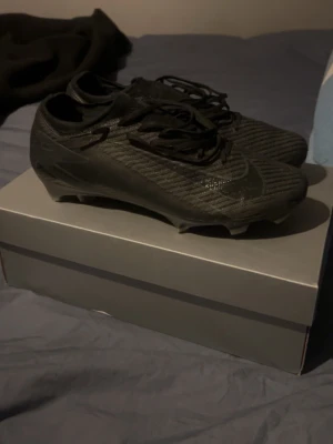 Svarta Nike fotbollsskor (elite)  - Säljer ett par helsvarta fotbollsskor från Nike med låg profil och snörning. Skorna har en modern, strömlinjeformad design och är tillverkade i syntetmaterial för bra grepp och kontroll på planen. Perfekta för dig som vill ha stilrena och funktionella skor till matchen.