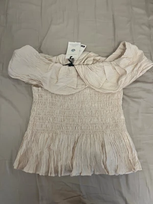 Beige offshoulder topp från Bik Bok - Supersöt beige offshoulder topp från Bik Bok med volangkant och smockad midja. Toppen har en luftig och skrynklig struktur som ger en avslappnad men ändå chic look. Perfekt till sommaren och lätt att styla med både jeans och kjol.