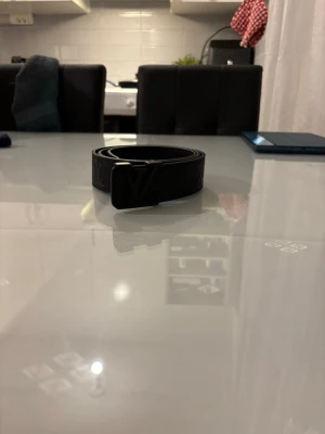 Svart skärp i skinn - Snyggt svart skärp med klassiskt spänne och präglat monogram längs hela bältet. Tillverkat i skinn och passar perfekt för att lyfta vilken outfit som helst. En riktig statement-accessoar för dig som gillar exklusiv stil. Den är 110 cm. Självklart aldrig använd. Tveka inte att höra av er vid frågor eller funderingar