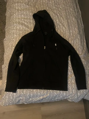 Svart hoodie från Polo Ralph Lauren - Snygg svart hoodie från Polo Ralph Lauren med dragkedja framtill och klassisk huva med dragsko. Liten vit logga broderad på bröstet. Tillverkad i mjukt bomullsmaterial som är skönt att ha på sig. Perfekt för en avslappnad och stilren look.
