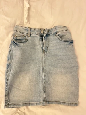 Ljusblå jeanskjol med fickor - Trendig ljusblå jeanskjol i kort modell med klassiska fem fickor, bälteshällor och knapp- samt dragkedjestängning framtill. Snygg tvättad denimlook som passar perfekt till sneakers eller boots.