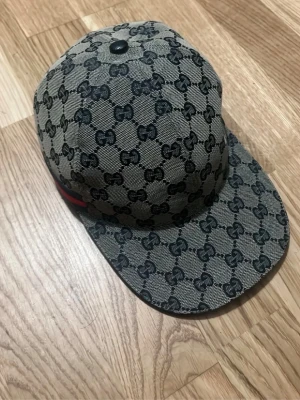 Gucci keps med GG-mönster - Snygg keps från Gucci med klassiskt GG-monogram i svart på beige bakgrund. Kepsen har böjd skärm och en diskret röd-grön rand på sidan. Tillverkad i canvasmaterial med mörkblått innerfoder. Perfekt för dig som vill ha en exklusiv och trendig accessoar.