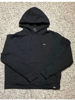 Svart hoodie från Dickies - Klassisk svart hoodie från Dickies med huva och dragsnören. Hoodien har en magficka framtill och ribbade muddar vid ärmslut och nederkant. Liten Dickies-logga på bröstet och vid nederkanten. Perfekt för en avslappnad streetwear-look.