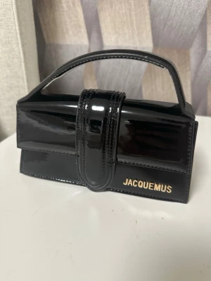 Jacquemus väska - Säljer en svart handväska i lack från Jacquemus med guldfärgad logga på framsidan. Väskan har ett stilrent, rektangulärt format och ett kort handtag. Stängs med ett brett lock och har guldfärgade metalldetaljer på sidorna. Perfekt för dig som gillar trendiga accessoarer.