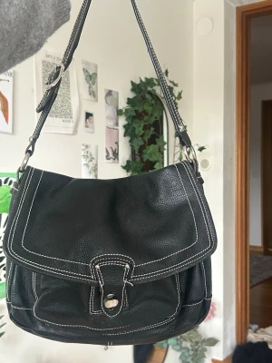 Svart axelremsväska med vita sömmar - Snygg svart handväska i mjukt läder med kontrasterande vita sömmar och silverfärgad knappstängning. Väskan har en bred axelrem och praktisk ficka framtill. Perfekt för dig som vill ha en stilren och klassisk accessoar.