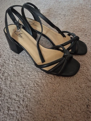 Svarta sandaler med klack från Clou - Stilrena svarta sandaler från Clou med fyrkantig tå, knutdetalj framtill och smala remmar runt ankeln. Klackhöjd i blockmodell för extra komfort. Ovandelen är i konstläder och innersulan är beige. Perfekta för dig som gillar enkel men trendig design.