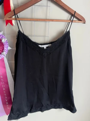Svart spetslinne från Kappahl - Säljer ett svart linne från Kappahl med tunna axelband och spetsdetaljer vid halsringning och nederkant. Perfekt att styla lager på lager eller bära som det är. Enkel och stilren design.
