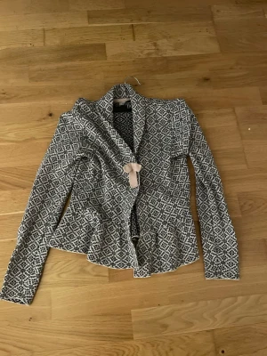 Mönstrad kofta med rosett - Supersnygg kofta från Odd Molly med svartvitt grafiskt mönster och en söt beige rosett framtill. Koftan har figurnära passform, lång ärm och volangkant nedtill. Perfekt för dig som gillar unika detaljer och skön stil. Aldrig använd i bra skick.