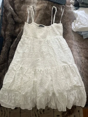 Vit sommarklänning med volanger - Söt vit kortklänning med smala axelband och volangkant. Klänningen har en smockad överdel och är tillverkad i ett lätt, strukturerat tyg som känns perfekt för varma dagar. Passformen är luftig och avslappnad, vilket ger en skön känsla.