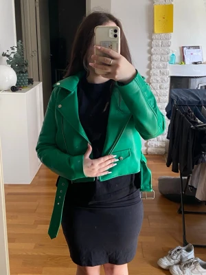 Grön skinnjacka med bälte - Snygg grön skinnjacka i bikerstil med silvriga dragkedjor och detaljer. Jackan har ett brett bälte i midjan och klassisk krage med tryckknappar. Perfekt statement-plagg för dig som vill sticka ut. Storlek S. Har katter hemma!!